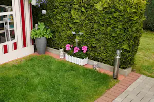 Terrasse am Campingplatz mit Hecke, Blumenbeeten und Pflanzkübeln neben dem Eingang, daneben gepflasterter Weg.