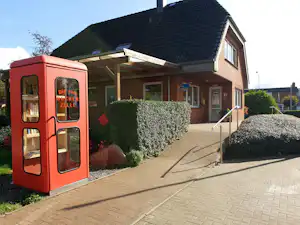 Roter Wohnwagen am Campingplatz neben einem Haus mit Eingang, gepflastertem Weg und Hecke.