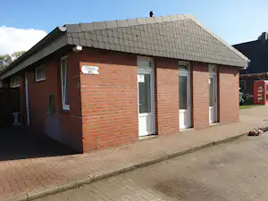 Ziegelhaus am Campingplatz mit Satteldach, weißen Fenstern und gepflastertem Weg; daneben ein rotes Gebäude und ein Fahrradständer.