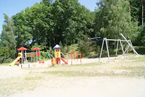 Spielplatz mit Schaukeln und Rutsche, gelben und roten Kletterelementen auf Sand, umgeben von hohen Bäumen.