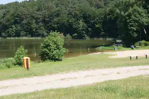 Seeufer am Campingplatz: grünes Gras, Sandweg, Bänke und ein Boot am ruhigen Wasser vor dichtem Wald.