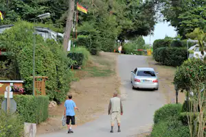 Campingplatz-Weg mit geparktem Auto, Wohnwagen und Zelten zwischen Büschen und Bäumen.