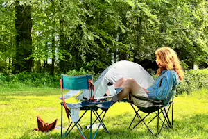 Frau sitzt im Campingstuhl am Klapptisch vor einem Zelt im Grünen, umgeben von Bäumen und Rasen.