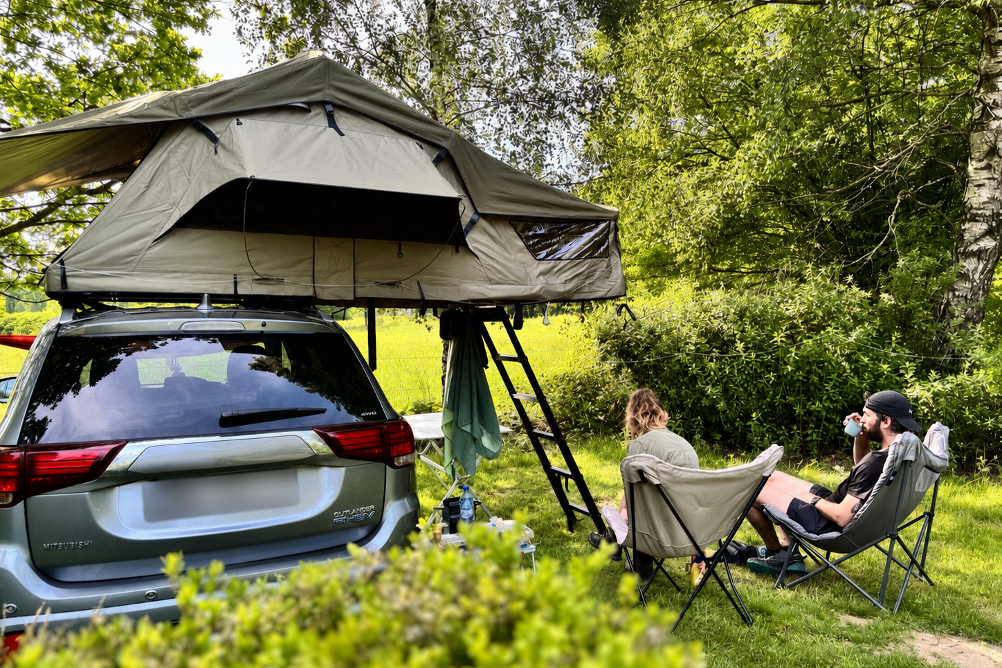 Camping am Auto: Zelt auf dem Dachgepäckträger, zwei Personen sitzen an Klappstühlen auf einer Wiese im Grünen.