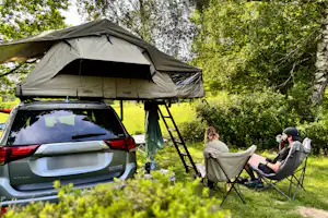 Camping am Auto: Zelt auf dem Dachgepäckträger, zwei Personen sitzen an Klappstühlen auf einer Wiese im Grünen.