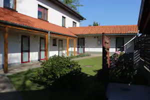 Reihenhausartige Ferienunterkunft mit Terrasse, Holzüberdachung, roten Türen und gepflegtem Garten mit Blumen am Campingplatz