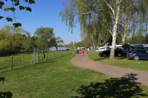 Campingplatz-Allee am See mit Rasen, Bäumen, Kiesweg und geparkten Autos, im Hintergrund Wohnwagen und Zelte.