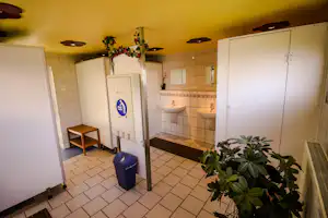 Sanitärbereich im Camping-Badezimmer mit Waschbecken, Toiletten, Fliesenboden und Abfalleimer, daneben Pflanze im Eingangsbereich.