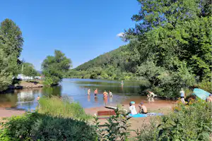 Flussufer mit Badebereich und Liegestühlen, Menschen im Wasser, umgeben von Bäumen und grünen Hügeln.