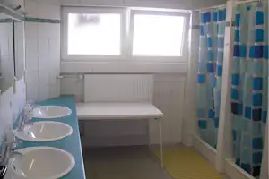 Waschraum mit drei Waschbecken, Fenster und Heizkörper, gefliesten Wänden sowie Duschvorhang im Campingbad.