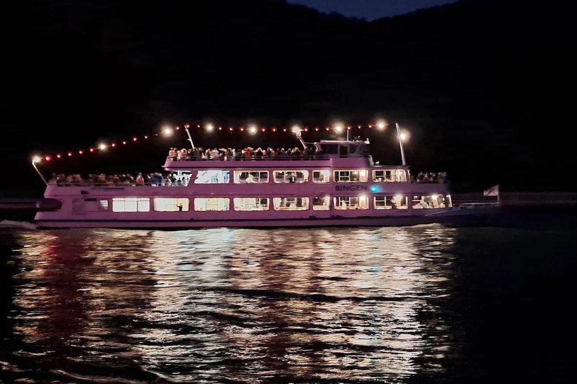 Ein beleuchtetes Ausflugsschiff fährt am Ufer entlang, reflektiert im Wasser, im Hintergrund dunkle Hügel und Uferlichter.