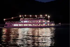 Ein beleuchtetes Ausflugsschiff fährt am Ufer entlang, reflektiert im Wasser, im Hintergrund dunkle Hügel und Uferlichter.