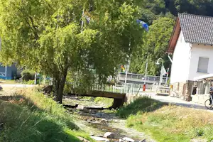 Bachlauf am Campingplatz mit Steinen und Gras, im Hintergrund Gebäude und Terrasse am Haus, umgeben von Bäumen.