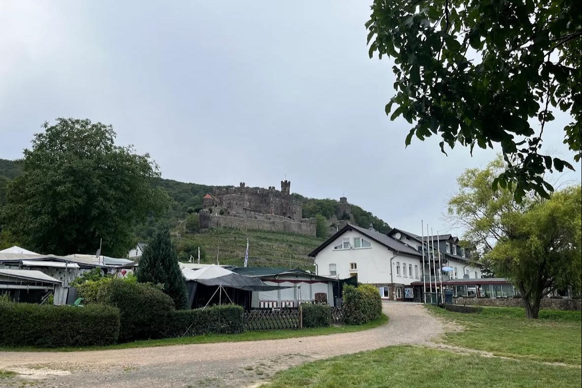 Campingplatz mit Kiesweg, Wohnwagen und Zelten, Hecken und Bäumen; im Hintergrund eine Burg auf dem Hügel.