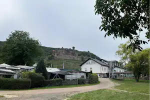 Campingplatz mit Kiesweg, Wohnwagen und Zelten, Hecken und Bäumen; im Hintergrund eine Burg auf dem Hügel.