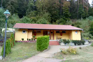 Ferienhaus mit rotem Ziegeldach, Eingang und Fenstern, davor gepflasterter Weg, umgeben von Büschen und Wald.