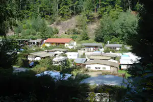 Campingplatz mit Wohnwagen und Zelten, kleinen Gebäuden und überdachten Stellplätzen im Grünen, umgeben von Bäumen am Hang.