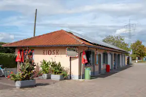 Kiosk-Gebäude mit roten Ziegeldach und Solarpanelen, Eingang mit Schiebetüren, Blumenbeete und Gehweg im Campingbereich.