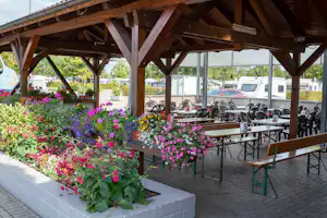 Blumenbeete und Terrasse unter einer Holzpergola mit vielen Stühlen und Tischen, im Hintergrund Wohnmobile und Zelte.