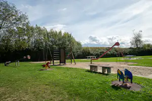 Spielplatz mit Klettergerüst und Rutsche, Sandfläche, Bänken und Spielgeräten im Grünen vor Bäumen.