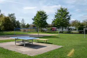 Tischtennis-Tisch auf gepflasterter Terrasse, dahinter Sandfläche, Zaun und Bäume, im Hintergrund Wohnmobile und Lieferwagen.