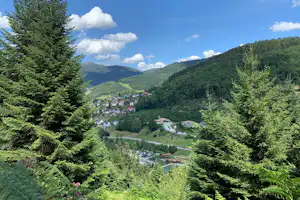 Blick ins Tal mit Campinganlage, umgeben von Nadelbäumen und grünen Hügeln, im Hintergrund Häuser und Wege.