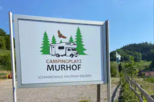 Wegweiser am Campingplatz Murhof mit Wohnmobil-Illustration, Schriftzug und Blick auf den Kiesweg zwischen Wiesen und Hügeln.