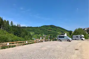 Stellplatz mit Wohnmobilen auf Schotterfläche, Holzgeländer und Blick auf grüne Hügel und Häuser im Tal.