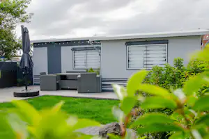 Wohnwagen mit Terrasse und Sitzbank, umgeben von grünem Gras und Büschen, im Hintergrund ein Gebäude mit Fenstern.