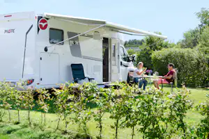 Wohnmobil mit Markise am Campingplatz, davor Garten mit Reben und zwei Personen am Tisch.