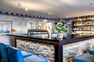 Blaue Sitzbank an der Bar-Theke mit Flaschenregal, Edelstahlspüle und Blumen auf dem Tresen im Innenbereich.