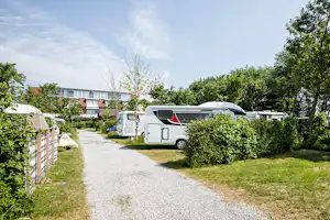 Wohnwagen auf einem Campingstellplatz, Kiesweg und Rasen zwischen Büschen und Bäumen, im Hintergrund ein mehrstöckiges Gebäude.