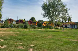 Bunte Fasssaunen und kleine Holzunterkünfte auf einer Wiese, umgeben von Büschen und Bäumen, im Hintergrund Gebäude.