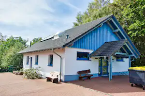 Blaues Ferienhaus mit Terrasse, Sitzbank und Mülltonnen am gepflasterten Weg, umgeben von Bäumen und Sträuchern.