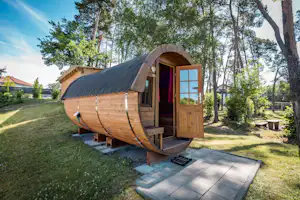 Glamping-Fasszelt mit offener Holztür, Terrasse und Holzbank auf einem gepflegten Campingplatz im Grünen, umgeben von Bäumen.