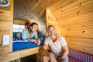 Zwei Personen sitzen in einer Holzunterkunft auf dem Bett, mit blauem Bettzeug und rotem Karomuster an der Wand.