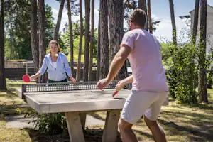 Zwei Personen spielen Tischtennis an einem Camping-Tisch im Grünen zwischen Bäumen und Hecken.
