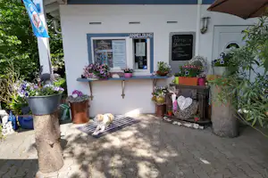 Eingang am Campingplatz mit weißen Wänden, Aushangtafel, Blumenampeln und Sitzbank vor dem Gebäude.