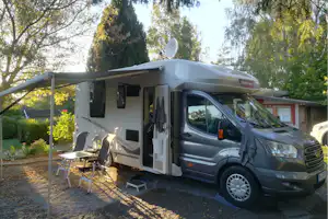 Wohnmobil mit Markise und Vorzelt auf einem Campingplatz, daneben Campingstühle und Tisch vor Bäumen.
