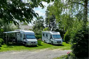 Zwei Wohnmobile auf einem Kiesplatz im Grünen, umgeben von Bäumen und Hecken, mit Rasenfläche am Stellplatz.