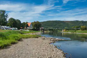 Campingplatz am Fluss mit Kiesufer, Brücke und Wohnwagen, im Hintergrund Häuser und bewaldete Hügel.