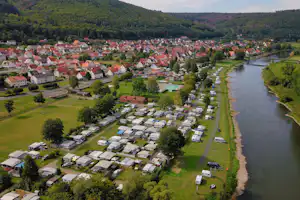 Luftaufnahme eines Campingplatzes mit Wohnwagen und Stellplätzen am Fluss, umgeben von Wiesen, Bäumen und einem Dorf.