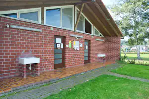 Ziegelhaus am Campingplatz mit Eingangstür, Fenstern, Waschbecken und gepflasterter Terrasse vor grünem Rasen.