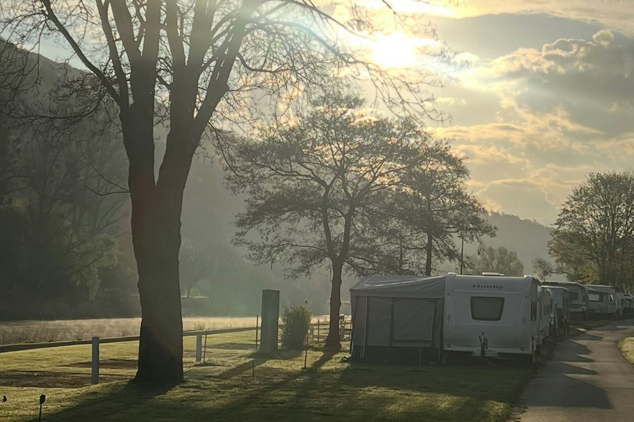 Wohnwagen auf einem Campingplatz, Reihe entlang eines Weges, umgeben von Bäumen und Wiese bei tiefstehender Sonne.