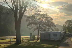 Wohnwagen auf einem Campingplatz, Reihe entlang eines Weges, umgeben von Bäumen und Wiese bei tiefstehender Sonne.