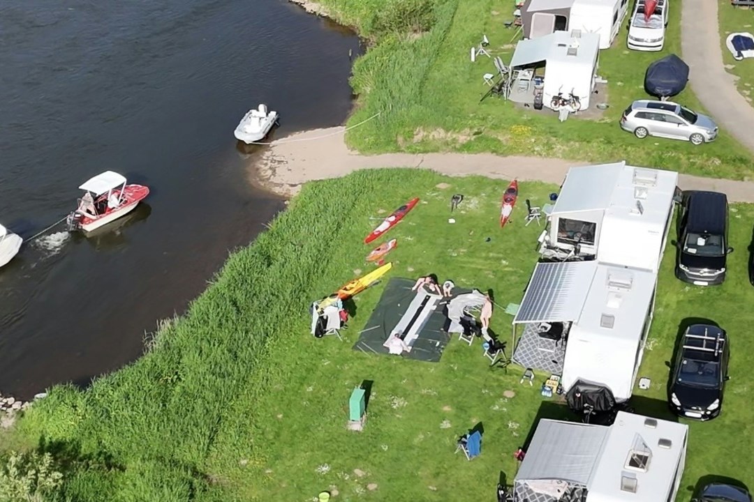 Campingplatz am Flussufer mit Wohnwagen und Autos, Rasenfläche, Booten im Wasser und Stegen am Ufer.