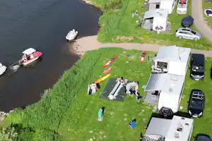 Campingplatz am Flussufer mit Wohnwagen und Autos, Rasenfläche, Booten im Wasser und Stegen am Ufer.