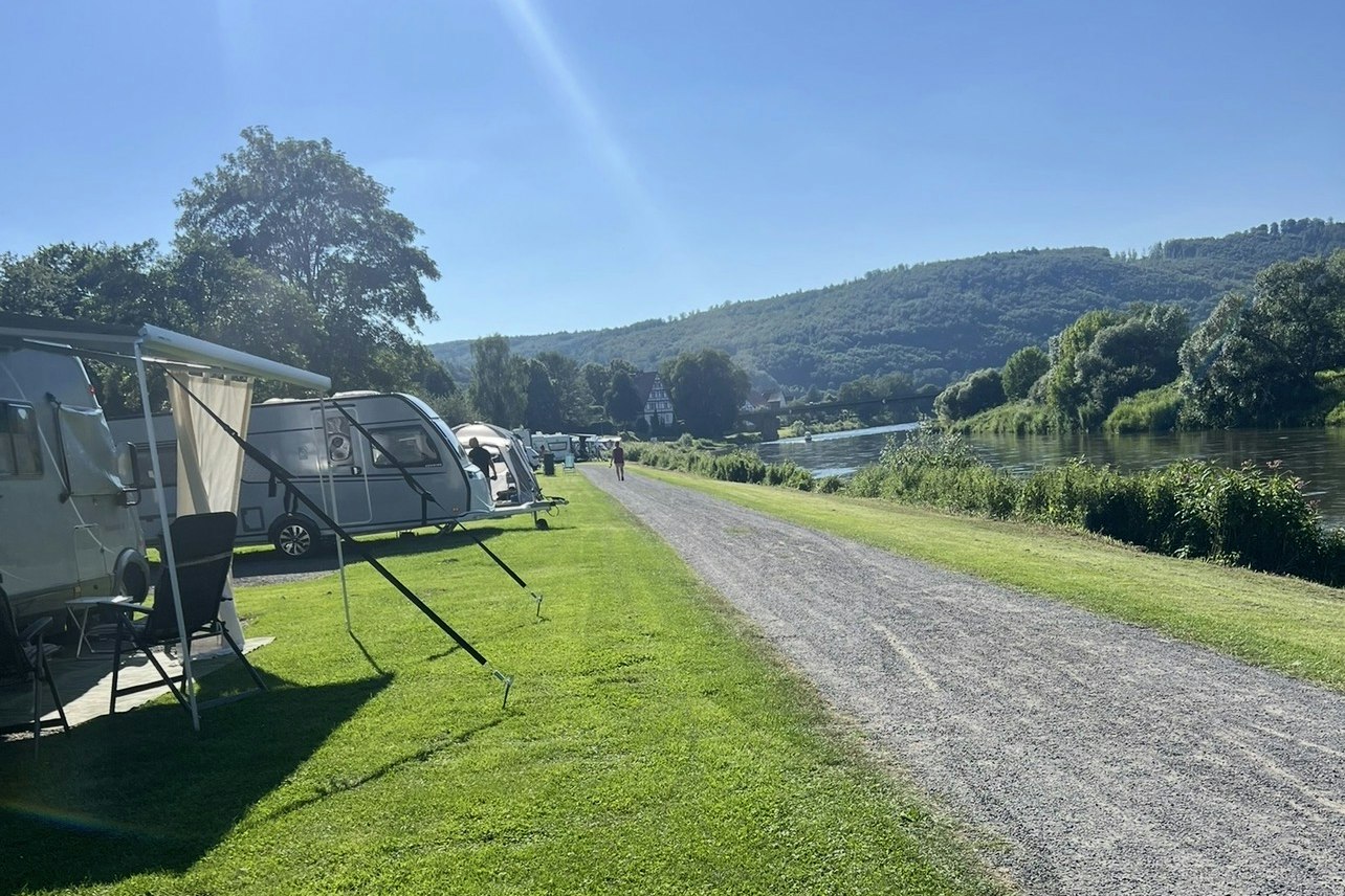 Stellplatz am Campingplatz mit Wohnwagen und Vorzelt, Rasenfläche und Kiesweg entlang eines Sees, im Hintergrund grüne Hügel.