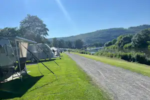 Stellplatz am Campingplatz mit Wohnwagen und Vorzelt, Rasenfläche und Kiesweg entlang eines Sees, im Hintergrund grüne Hügel.