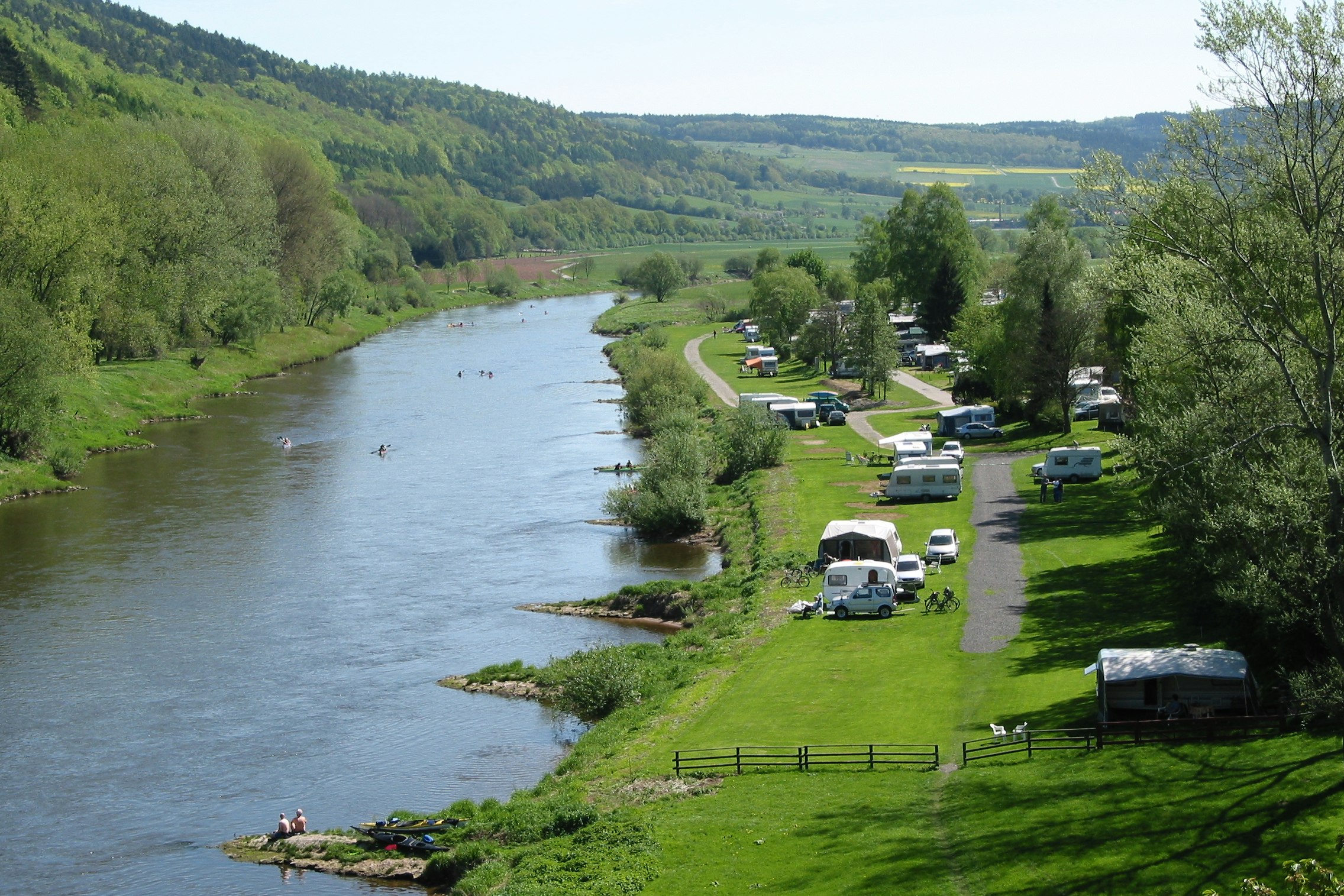 Campingplatz am Fluss mit Wohnmobilen und Stellplätzen auf der Wiese, Blick auf das Ufer und die grünen Hügel.